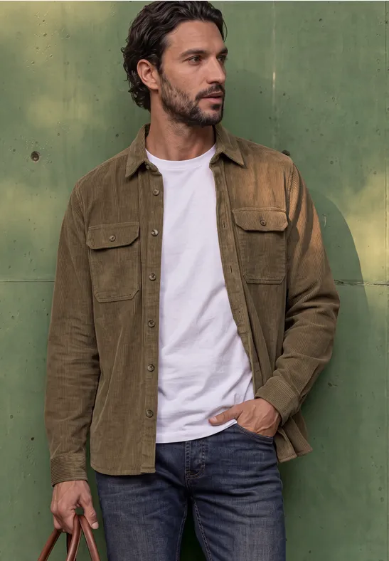 Ein Mann in weißem T-Shirt, brauner Jacke und Jeans steht vor grüner Wand und blickt zur Seite.