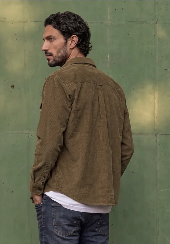 Ein Mann mit brauner Jacke und Jeans blickt über die Schulter vor einer grünen Wand.