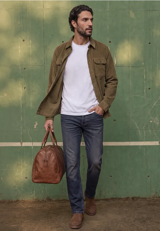 Ein Mann lehnt lässig vor grüner Wand, trägt braune Jacke, weißes T-Shirt, Jeans und hält eine Lederreisetasche.