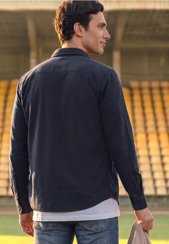 Ein Mann im dunklen Hemd steht auf einem Fußballfeld mit leeren Tribünen im Hintergrund.