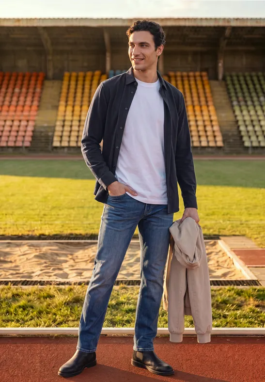 Ein lässig gekleideter Mann mit Jeans und Jacke steht neben einer Weitsprunggrube auf einer Sportanlage.