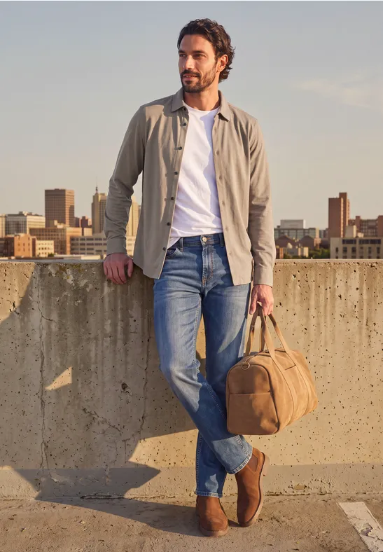 Ein lässig gekleideter Mann lehnt auf einem Parkdeck mit Tasche, im Hintergrund Stadtpanorama.