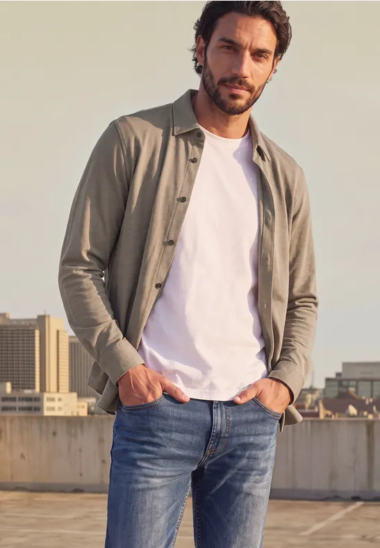 Ein bärtiger Mann im weißen T-Shirt und offenem Hemd auf einem Dach vor Stadtblick.
