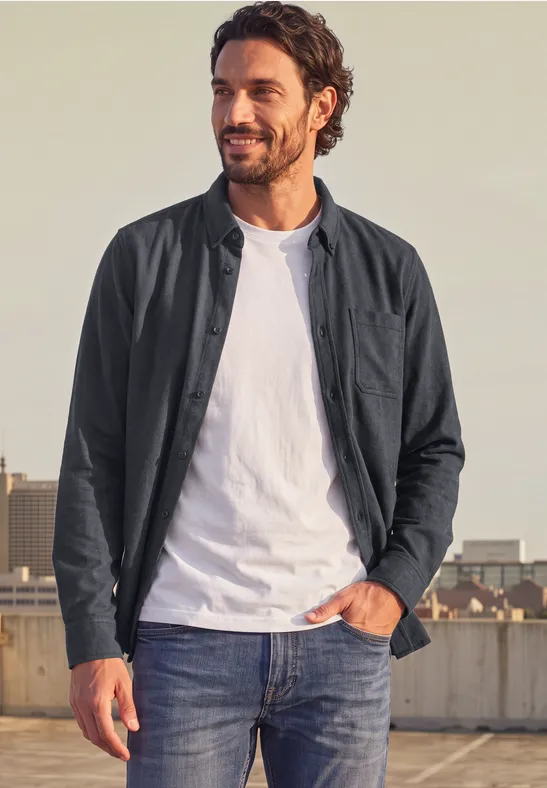 Mann steht auf Dachterrasse, trägt Jeans, weißes T-Shirt und dunkle Jacke, lächelt Richtung Kamera.