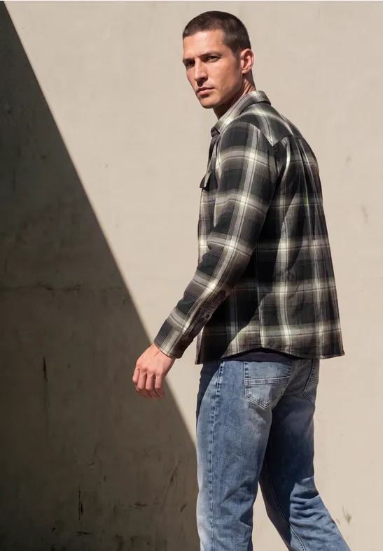 Ein Mann in kariertem Hemd und Jeans läuft vorbei und schaut über die Schulter vor einer hellen Wand.