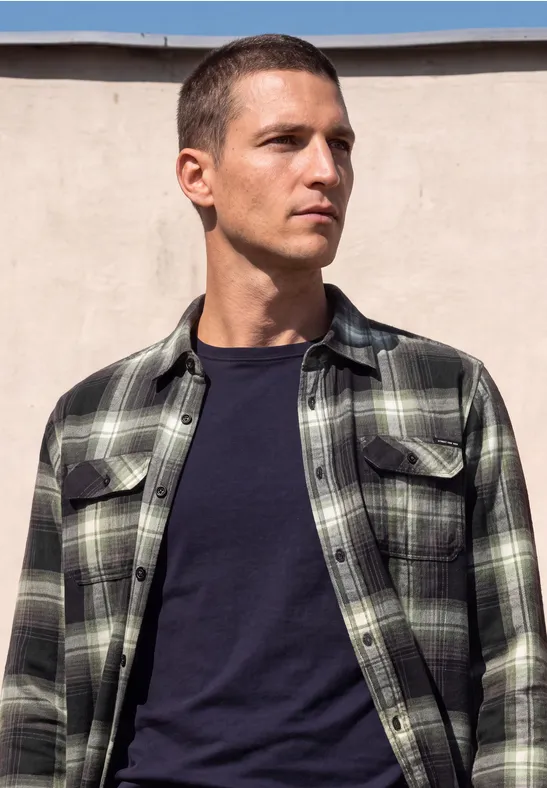 Ein Mann mit kurzem Haar trägt ein grünes kariertes Hemd über einem schwarzen T-Shirt vor einer Betonwand.