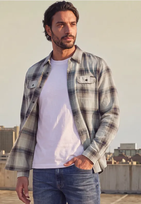 Ein Mann in kariertem Hemd und weißem T-Shirt steht vor urbaner Kulisse und schaut nach rechts.