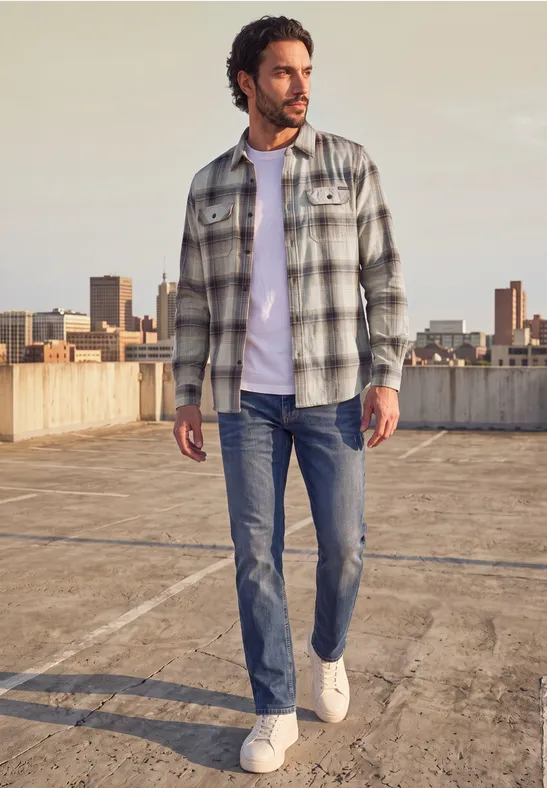 Ein Mann mit kariertem Hemd, weißem T-Shirt, Jeans und Turnschuhen steht auf einem Parkdeck mit Skyline im Hintergrund.
