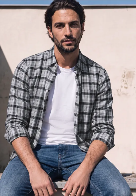 Ein Mann in karierter Hemdjacke, weißem T-Shirt und Jeans sitzt entspannt und schaut in die Kamera.