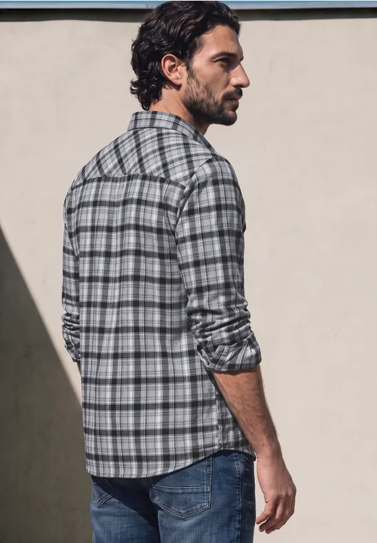 Ein Mann im karierten Hemd und Jeans steht mit dem Rücken zur Kamera vor einer neutralen Wand.