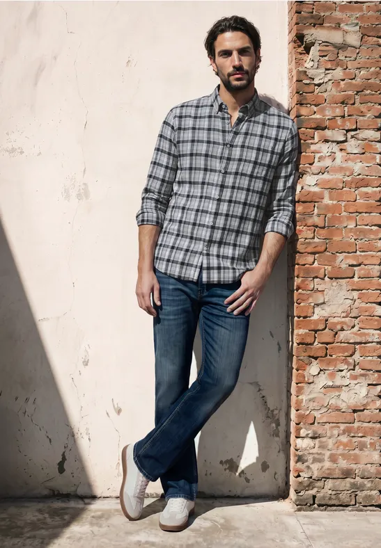 Ein Mann lehnt an einer Backsteinwand, trägt kariertes Hemd, Jeans und weiße Sneaker im Sonnenlicht.