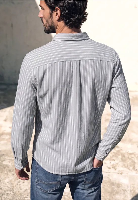 Ein Mann im hellblauen, gestreiften Hemd und Jeans steht mit dem Rücken zur Kamera vor einer grauen Wand.