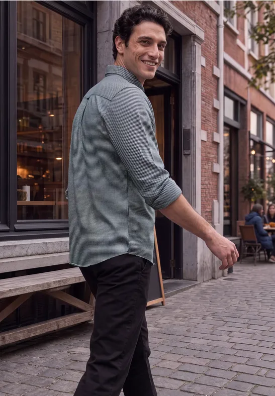 Un homme en chemise verte et pantalon noir marche en souriant sur une rue pavée devant un café.