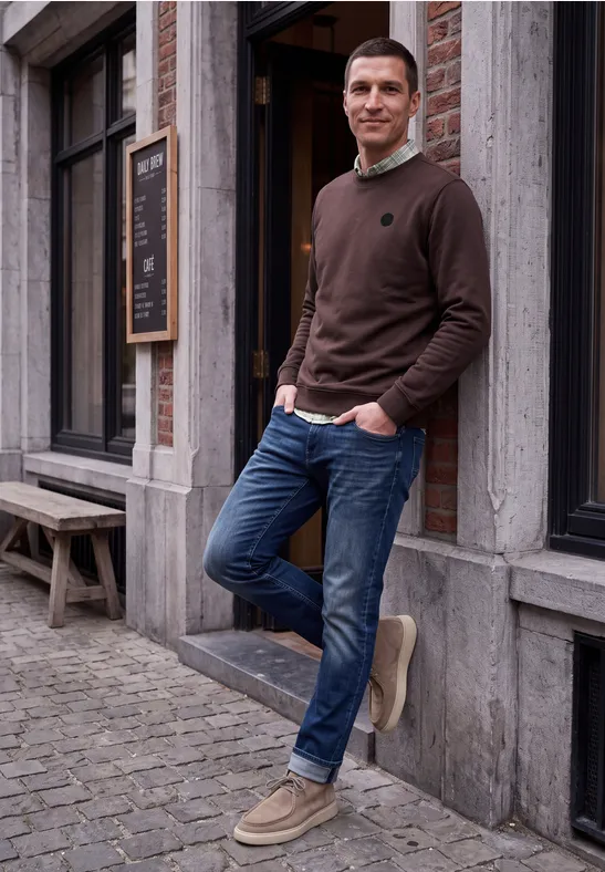 Ein lächelnder Mann in braunem Pulli, Jeans und Boots lehnt an einer Hauswand vor einem Geschäft.