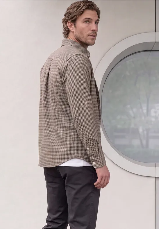 Un homme en chemise beige et pantalon foncé regarde par-dessus son épaule devant une fenêtre ronde.