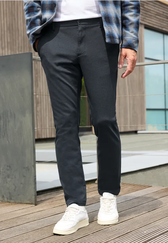 Een man draagt een slanke donkergrijze broek en witte sneakers op een houten terras voor een modern gebouw.