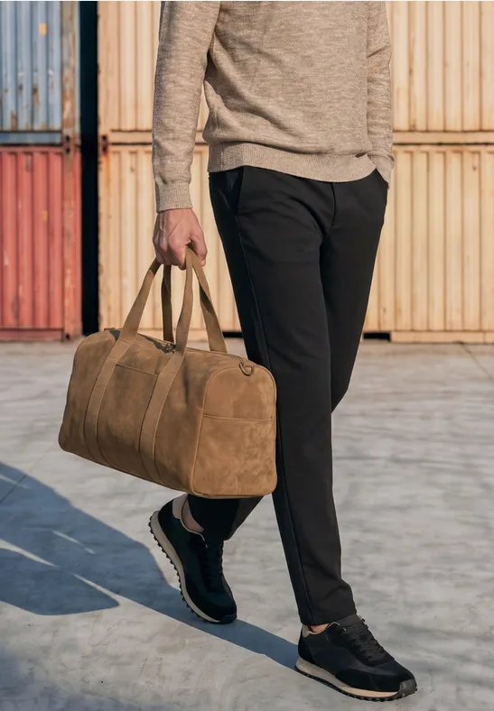 Eine Person trägt schwarze Hose und hält eine braune Reisetasche vor bunten Containern.