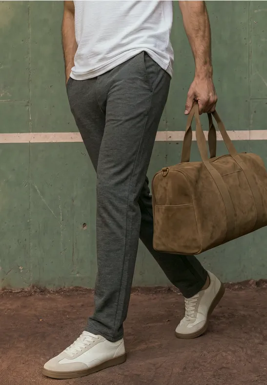 Ein Mann in grauen Hosen und weißen Sneakern hält eine braune Weekender-Tasche vor grünem Hintergrund.