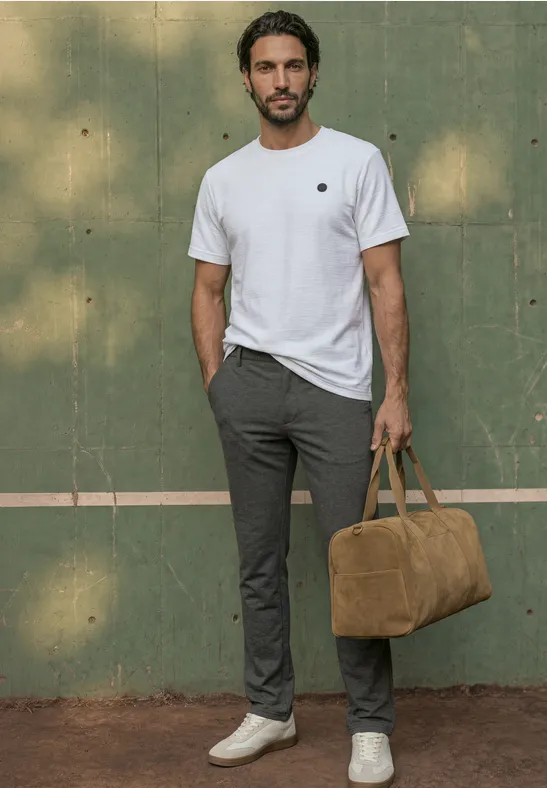 Ein Mann mit weißen Sneakers, grauen Hosen und weißem T-Shirt hält eine braune Tasche vor grünem Hintergrund.