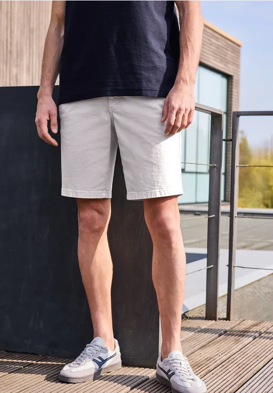 Mężczyzna w czarnej koszulce, szarych szortach i sneakersach, stojący na drewnianym tarasie przed nowoczesnym budynkiem.