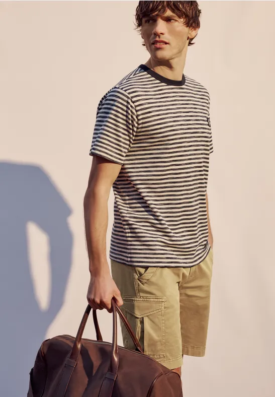 Een man draagt een gestreept T-shirt, kaki shorts en houdt een bruine leren tas in zijn hand.