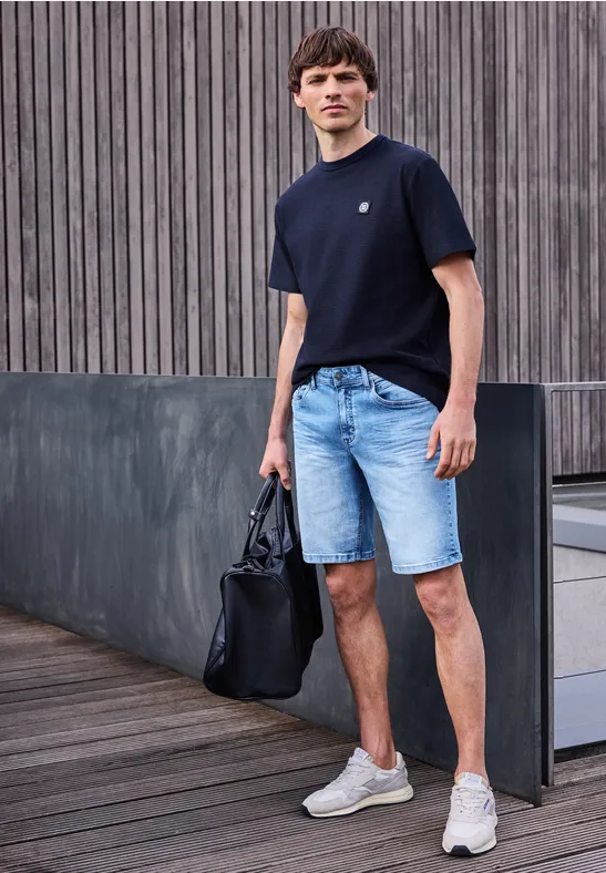 Ein Mann in lässigem Outfit: schwarzes T-Shirt, Jeans-Shorts und weiße Sneaker mit schwarzer Tasche auf Holzdeck