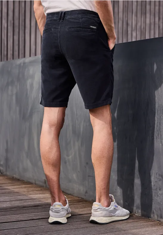 Ein Mann steht auf einer Holzplattform, trägt schwarze Jeans-Shorts und weiße Sneakers, Hände in den Taschen.