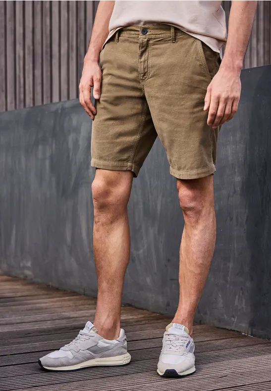 Ein Mann in khakifarbenen Shorts und grauen Sneakern steht vor einer Holz- und Betonwand.