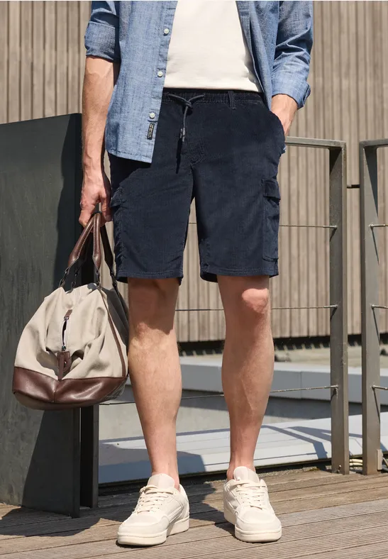 Man in denimoverhemd, wit T-shirt, donkerblauwe corduroyshort, witte sneakers en reistas.