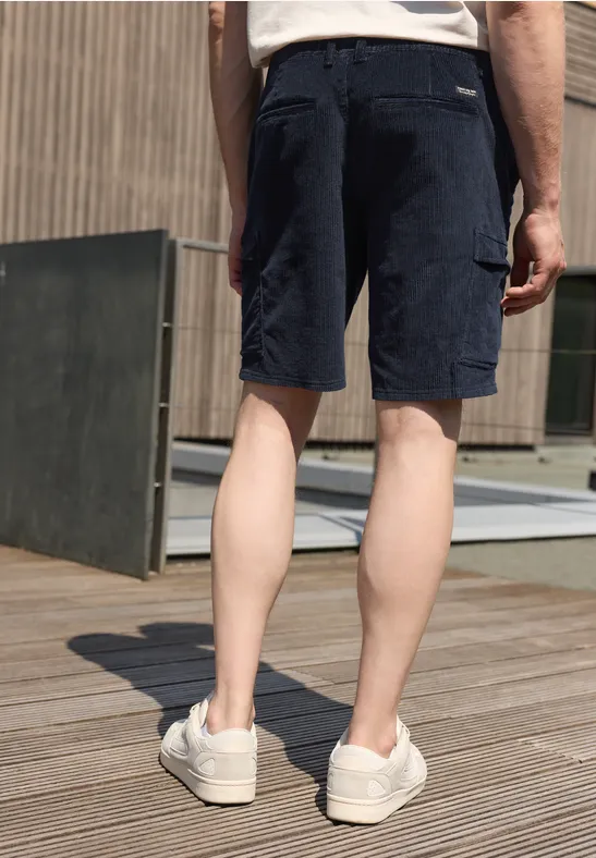 Man van achter gefotografeerd in marineblauwe cargoshorts en witte sneakers op houten dek.