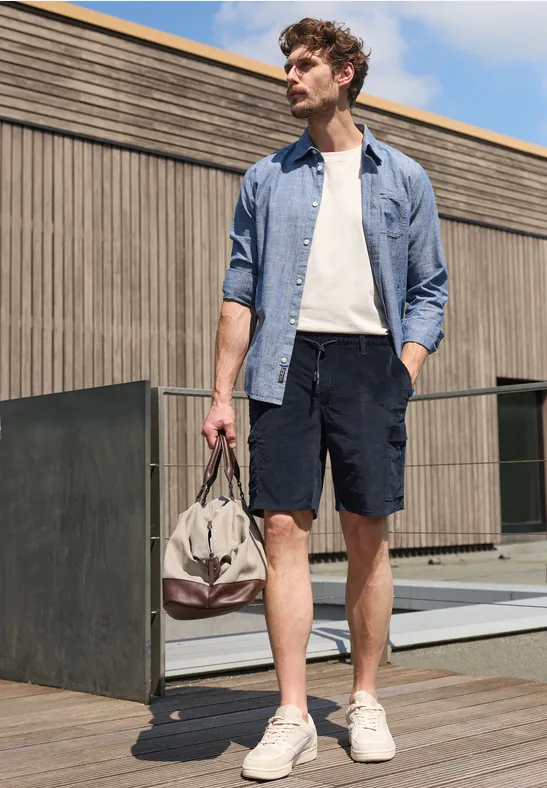 Een man in overhemd, t-shirt, korte broek en sneakers staat buiten en houdt een reistas vast.