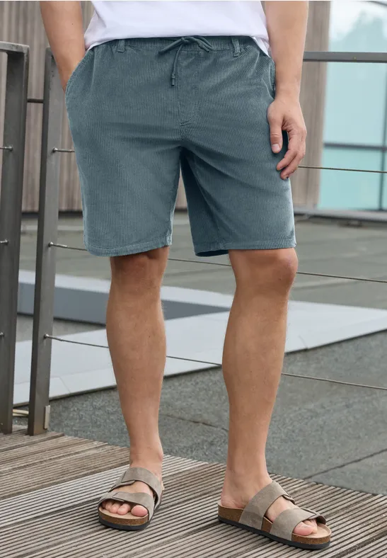 Mann steht auf Holzterrasse in grauen Shorts und Sandalen vor Glas- und Metallgeländer.