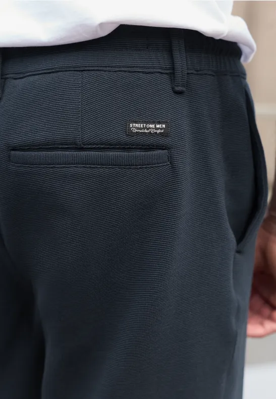 Nahaufnahme einer Person in dunkelblauer Hose mit Gesäßtasche und kleinem Label, weißes Shirt sichtbar.