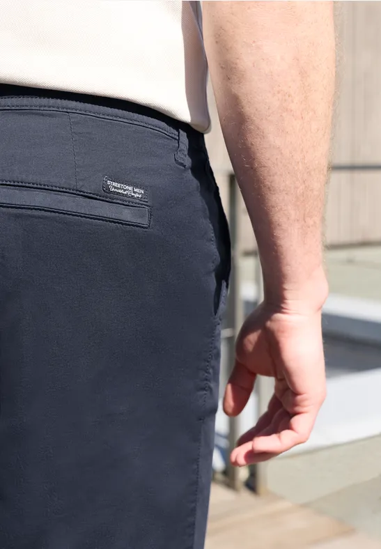 Rückansicht: Mann im Freien trägt dunkelblaue Hose mit Reißverschlusstasche, helles Oberteil und lockere Handhaltung.