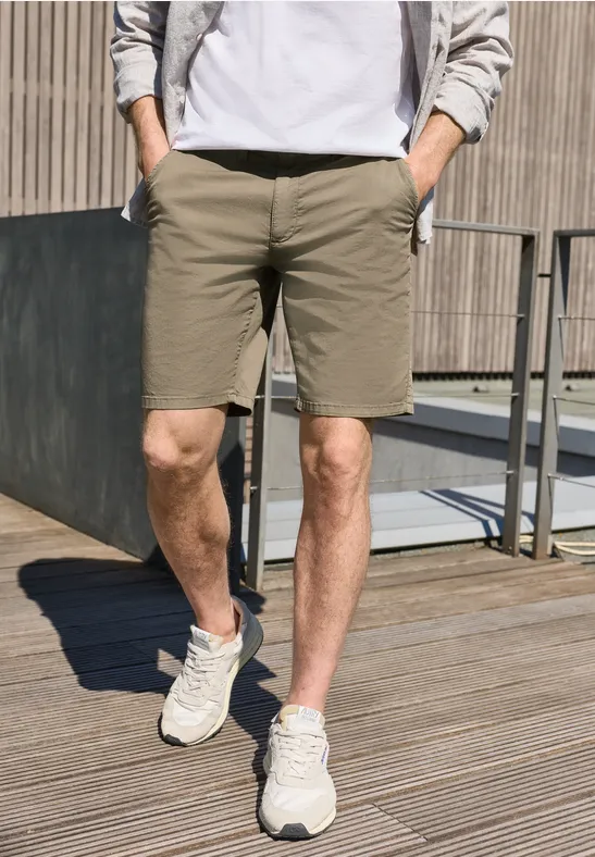 Ein Mann steht auf Holzdeck, trägt khakifarbene Shorts, weiße Sneaker und eine leichte Jacke.