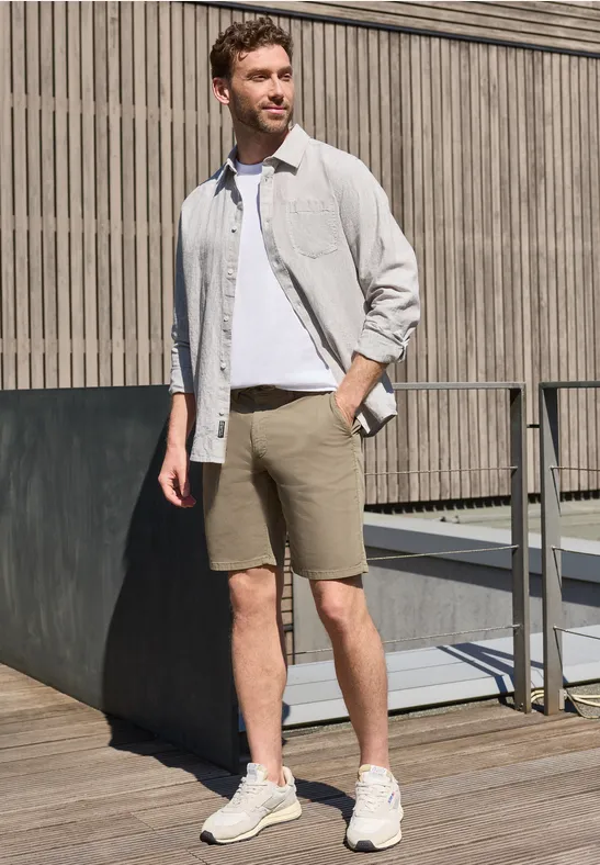 Ein Mann in weißem T-Shirt, hellgrauem Hemd, beigen Shorts und Sneakers steht auf einer Holzterrasse vor Holzpaneelen.