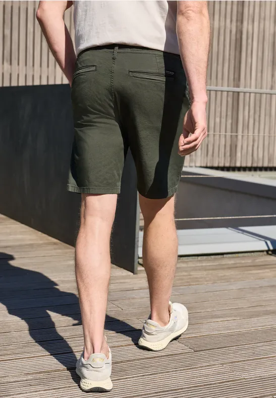Mann von hinten in dunkelgrünen Shorts und Sneakers, läuft über eine Holzterrasse an Gebäude.