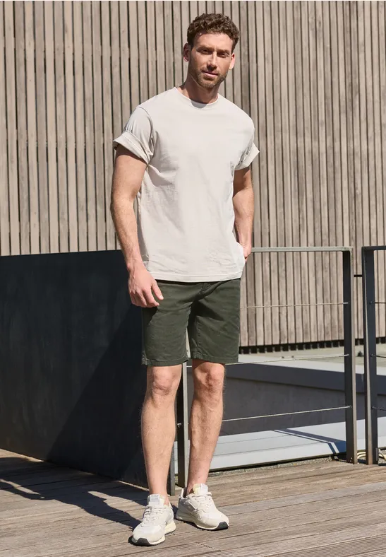 Ein Mann in weißem T-Shirt und grünen Shorts steht auf einer Holzterrasse neben einem Geländer.