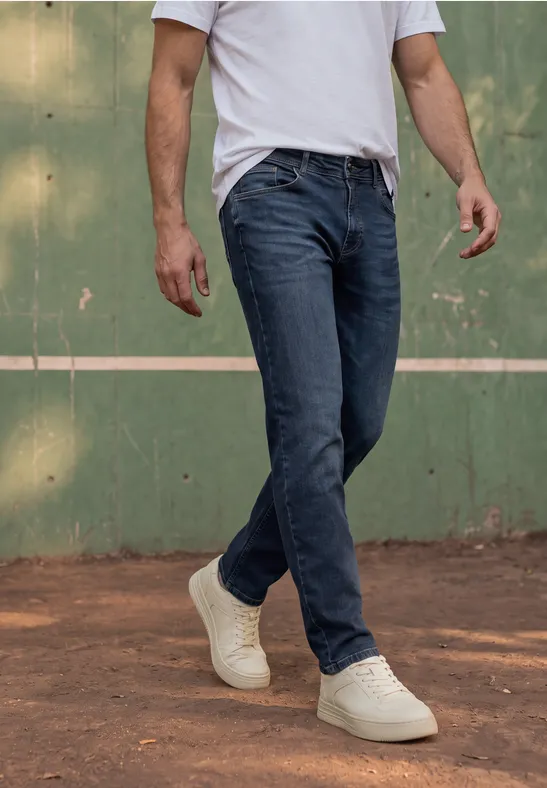 Un homme en jean, t-shirt blanc et baskets blanches marche sur un terrain de sport devant un mur vert.