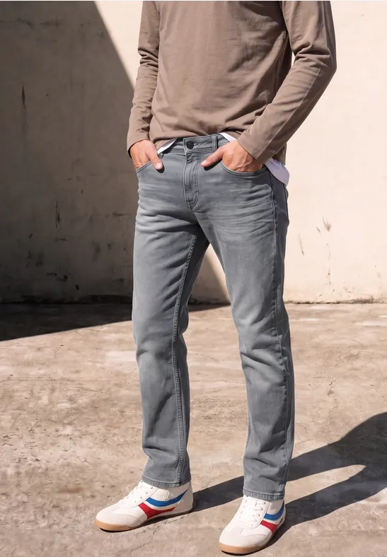 Homme en jean gris, haut à manches longues et baskets blanches à rayures rouge et bleu devant un mur.
