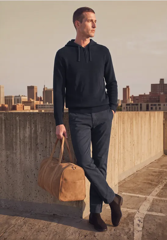 Man in casual kleding staat op een dak met stadszicht en houdt een bruine reistas vast.
