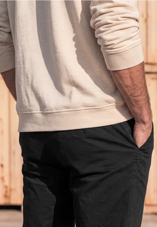 Ein Mann in beigem Pullover und schwarzer Hose steht seitlich vor einer Holzwand, die Hand in der Hosentasche.