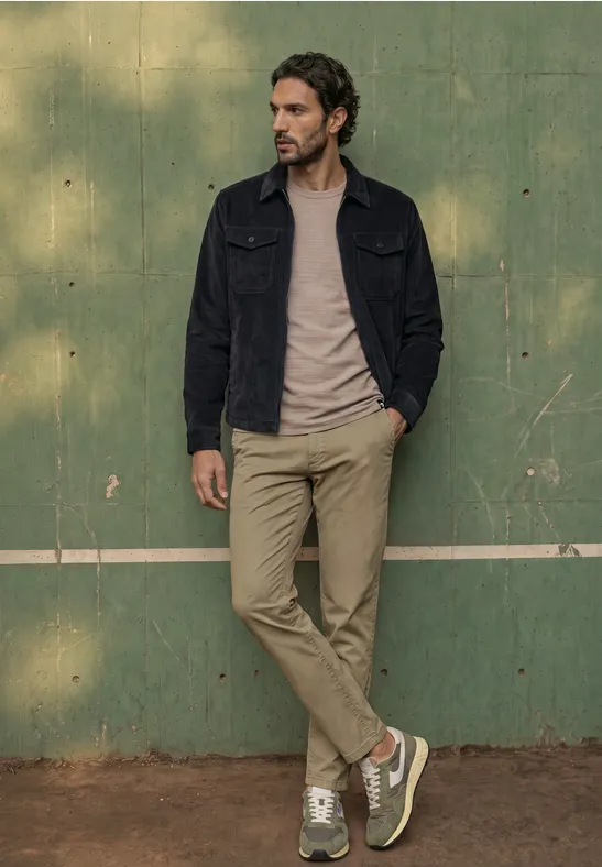 Ein bärtiger Mann lehnt lässig an einer grünen Wand, trägt Jeansjacke, T-Shirt und Sneakers.