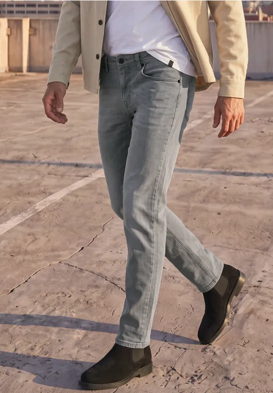 Ein Mann in grauer Jeans, beigem Sakko und schwarzen Stiefeln geht über einen Betonboden.