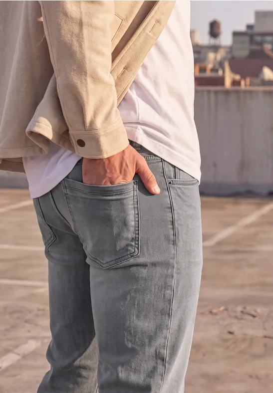 Mann steht auf Dach, trägt Jeans, weißes Shirt, beige Jacke, eine Hand in der Hosentasche.