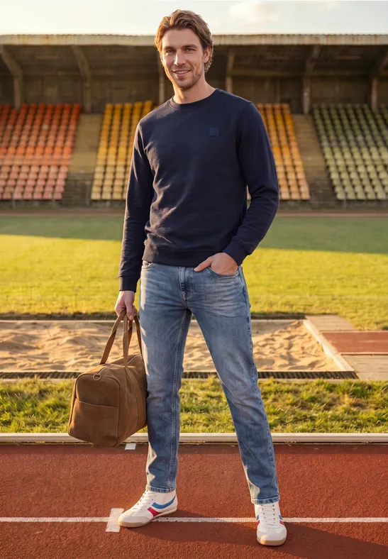 Een man staat op de atletiekbaan voor de verspringzandbak, houdt een tas vast, draagt jeans en trui.