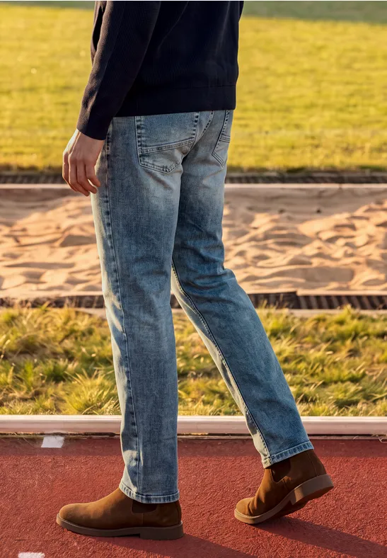 Eine Person in Jeans und braunen Schuhen geht auf einer Sportbahn neben einer Sandgrube.