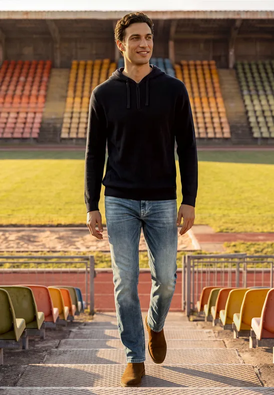 Ein Mann in Jeans und Kapuzenpullover geht in einem leeren Stadion auf den Tribünenweg zu.
