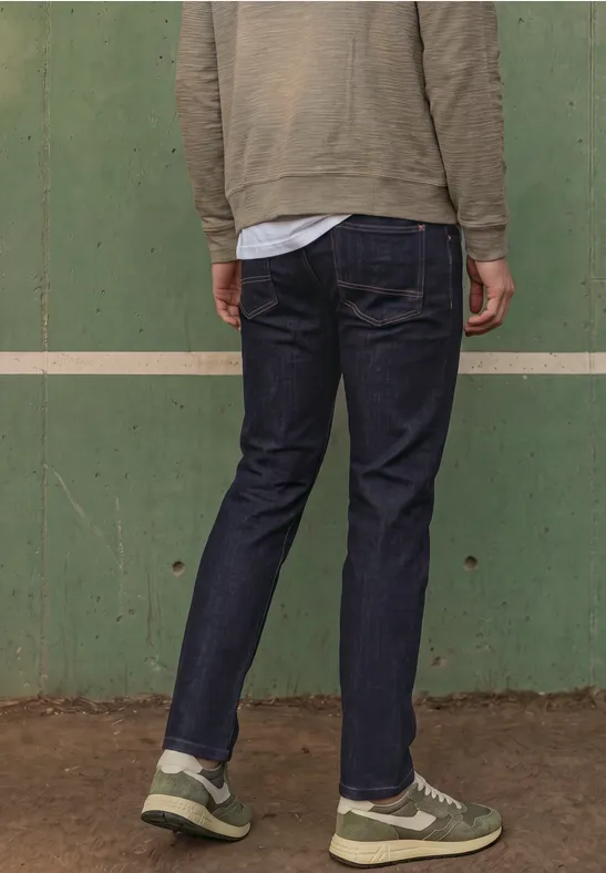 Person mit dem Rücken zur Kamera, trägt dunkle Jeans und Sneaker vor grüner Wand.