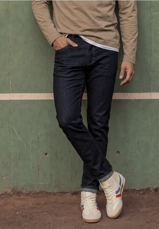 Ein Mann in Jeans und Sneakers steht vor einer grünen Wand, eine Hand in der Hosentasche.